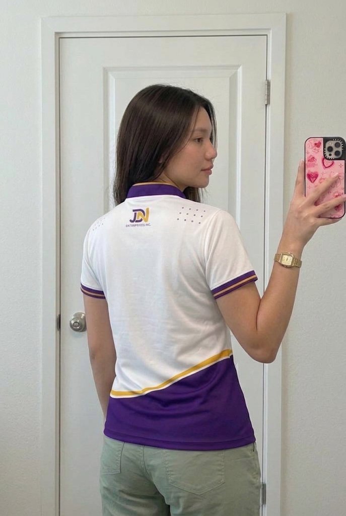 Team member showing the back of the JDN employee polo in a mirror: purple collar, JDN logo, purple wave with gold trim